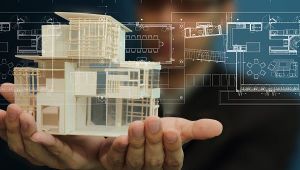 Två händer håller en modell av en byggnad, i bakgrunden syns en planritning