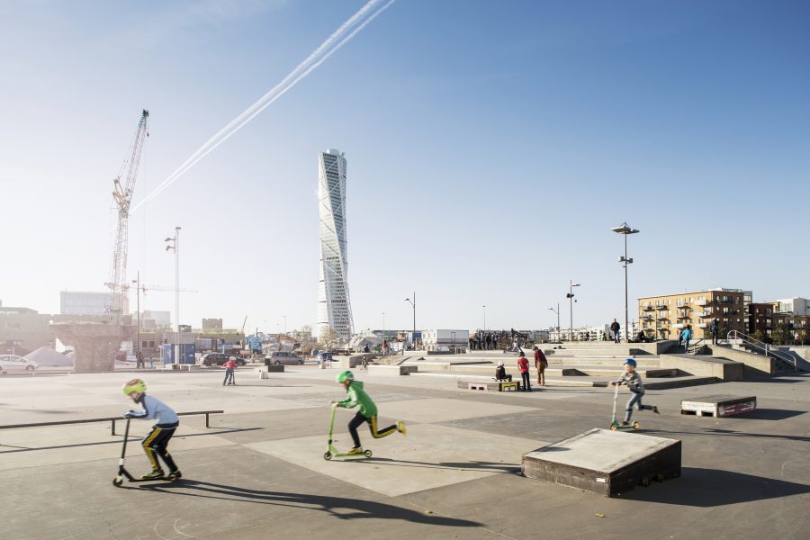 Barn åker sparkcykel och skateboard i en stadsmiljö