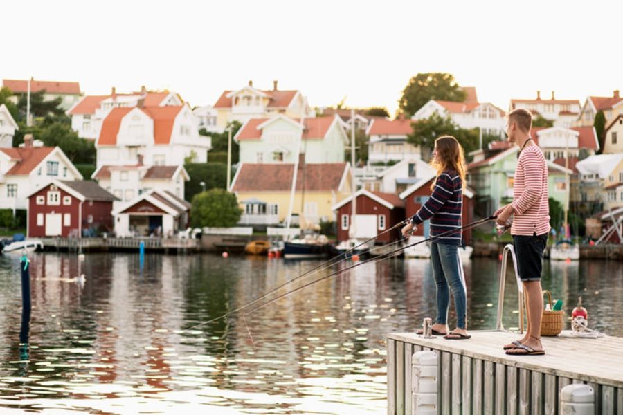 Två personer som står och fiskar på en brygga med hus i bakgrunden.