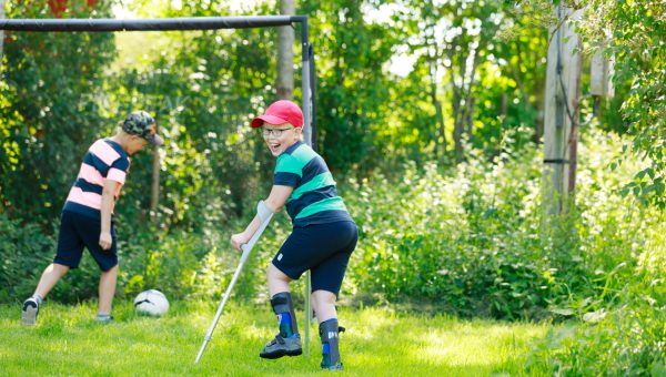 Två barn spelar fotboll på en gräsmatta. Ett barn har kryckor och springer med dem som stöd