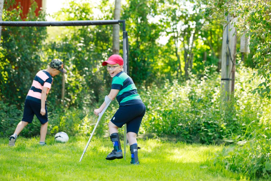 Två barn spelar fotboll på en gräsmatta. Ett barn har kryckor och springer med dem som stöd