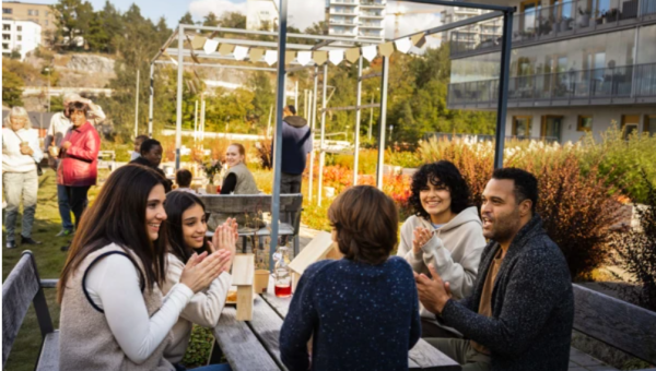 En grupp personer sitter utomhus vid ett picknickbord på innergården till ett flerbostadshus.